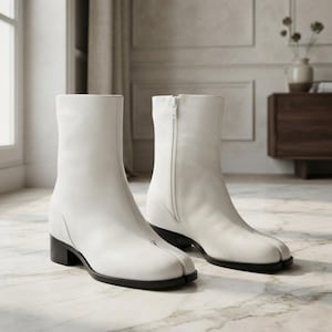 May include: A pair of white ankle boots featuring a distinct split-toe design, a side zipper closure, and a low block heel. The boots are constructed with a smooth finish and a structured silhouette, showcasing a clean and uniform white color throughout.