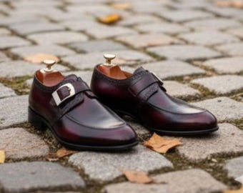Puede incluir: Un par de zapatos monje de cuero burdeos con hebillas plateadas. Los zapatos tienen un diseño clásico con punta redonda y están sobre una superficie adoquinada con hojas caídas.