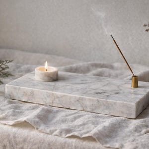 Könnte beinhalten: Ein rechteckiger Räucherstäbchenhalter aus Marmor mit einer brennenden Kerze und einem Messing-Räucherstäbchenhalter. Die Kerze und der Räucherstäbchenhalter sind auf der Marmoroberfläche platziert. Eine kleine Vase mit einem Zweig Grünzeug befindet sich im Hintergrund.