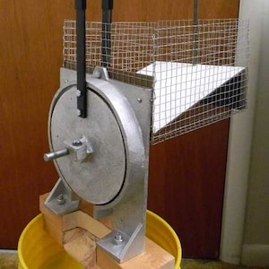 May include: A metal and wood device with a large circular wheel, two black handles, and a wire mesh enclosure. The device is mounted on a wooden base and a yellow bucket.