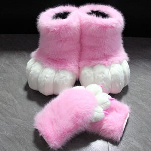 May include: A pair of ankle-high boots and matching fingerless gloves covered in soft pink faux fur. The boots and gloves feature white faux fur toe and finger pads at the ends, with small, pointed pink plastic claws attached to each digit.