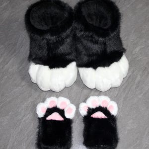 May include: A pair of black and white furry paw slippers and matching hand paws. The slippers have a black upper and white paw pads. The hand paws are black with white and pink details. The items are designed to resemble animal paws.