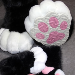 May include: Close-up of furry black and white paw pads. The pads have a white, fluffy exterior and a gray sole with pink paw prints. The image shows multiple paws, suggesting a costume or plush toy.