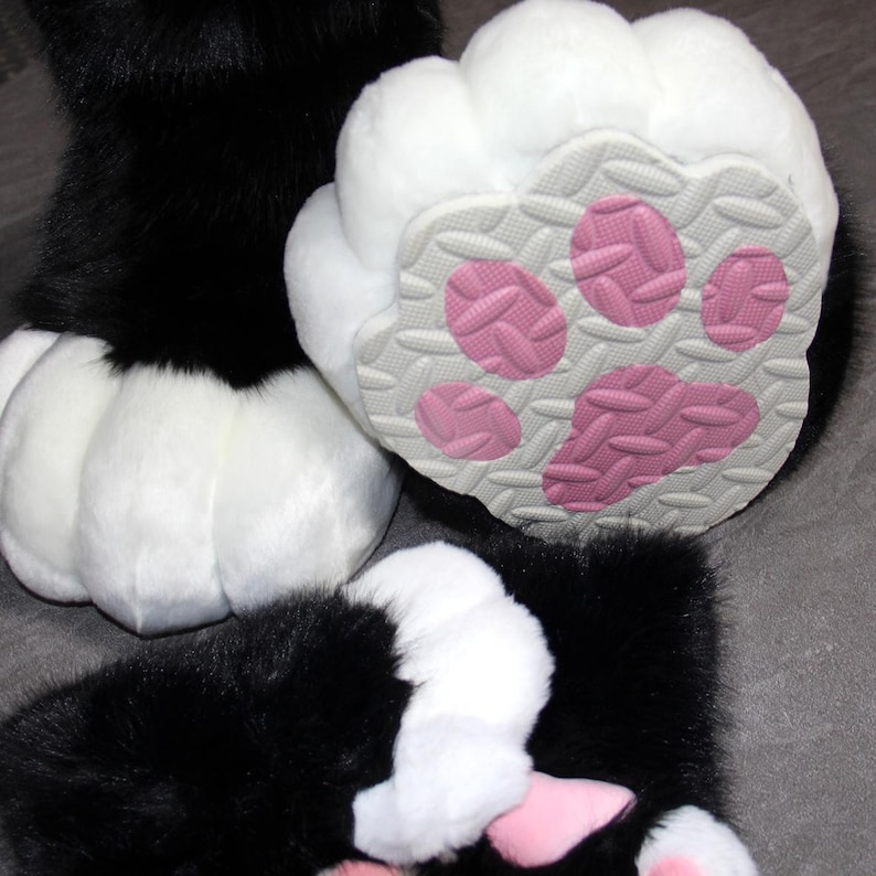 May include: Close-up of furry black and white paw pads. The pads have a white, fluffy exterior and a gray sole with pink paw prints. The image shows multiple paws, suggesting a costume or plush toy.