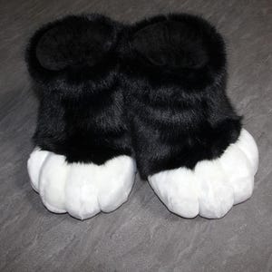May include: A pair of plush, black and white furry paw slippers. The slippers have large, white paw pads and black fur covering the upper portion. The slippers are designed to resemble animal paws.