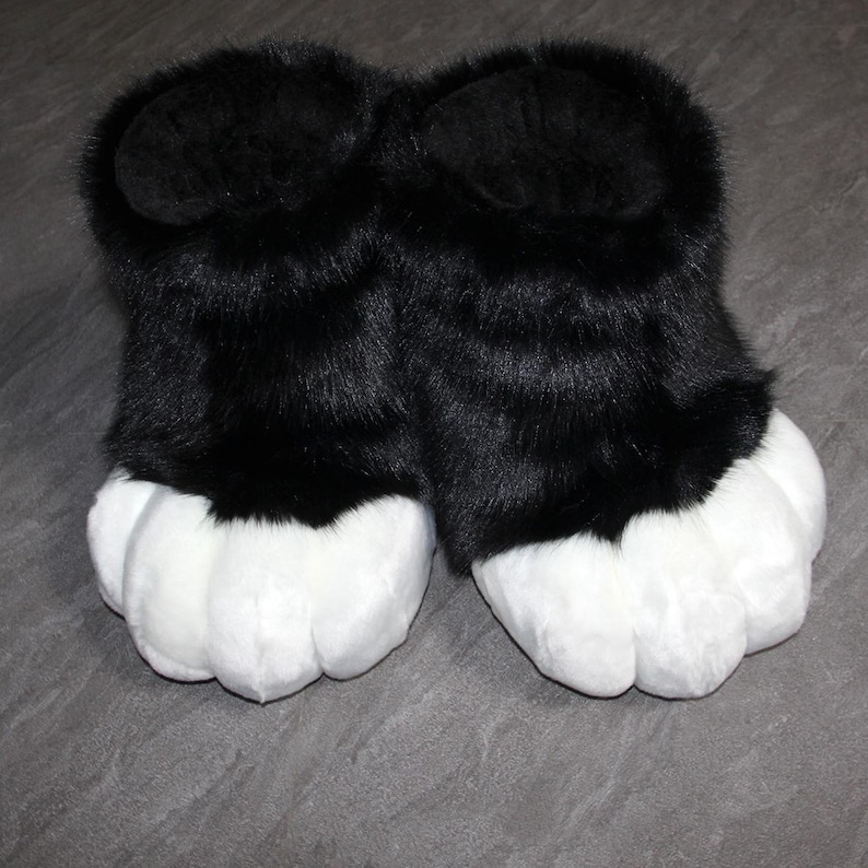 May include: A pair of plush, black and white furry paw slippers. The slippers have large, white paw pads and black fur covering the upper portion. The slippers are designed to resemble animal paws.