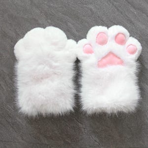 Peut inclure: Une paire de gants en fausse fourrure blanche en forme de pattes d'animal. Un gant montre le dos blanc et duveteux, tandis que l'autre présente la paume avec quatre coussinets roses arrondis et un grand coussinet central en forme de patte.