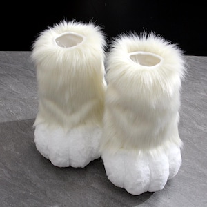 May include: A pair of ankle-high boot covers featuring long, cream-colored faux fur on the upper section and a shorter, dense white plush texture on the lower foot area, designed with a rounded, paw-like shape at the toe and an open top.