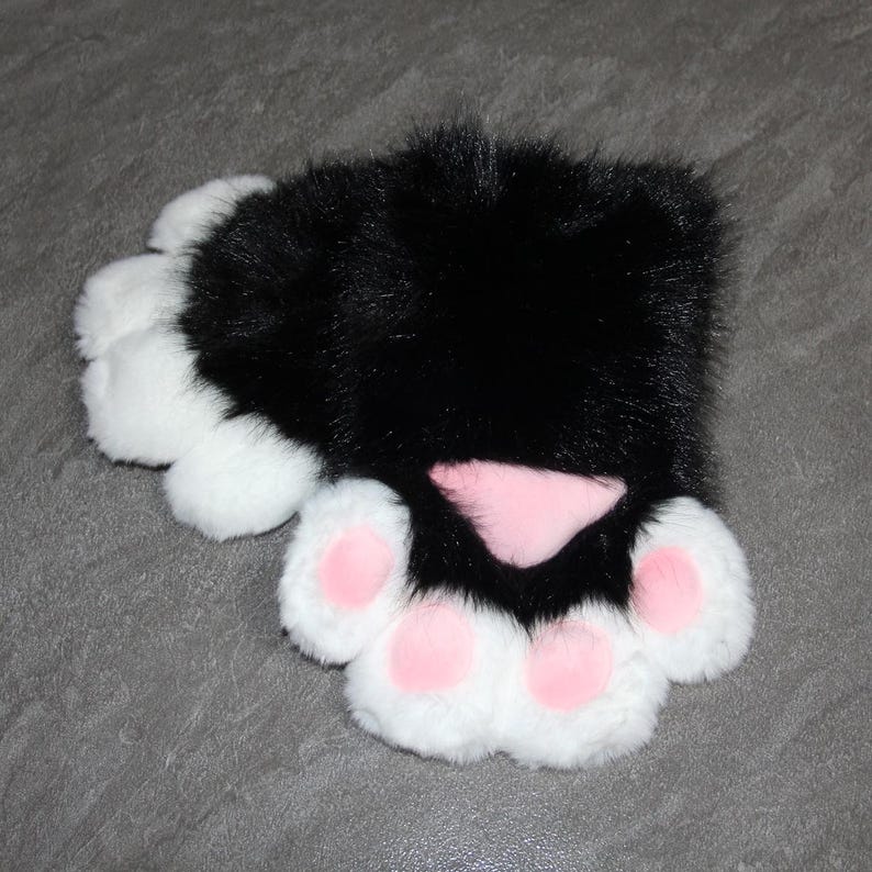 May include: A black and white faux fur paw pad with pink paw pads and a pink nose. The paw pad is designed to resemble a cat's paw. The product is likely a costume accessory or part of a plush toy.