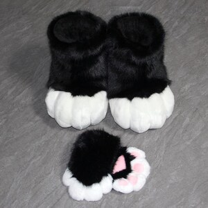 May include: A pair of black and white furry paw-shaped slippers with matching paw-shaped hand covers. The slippers and hand covers have a black upper portion and white paw pads. The hand covers have pink paw pads.