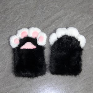 May include: A pair of black and white faux fur mittens shaped like cat paws. The mittens have white fur on the paw pads and pink accents. The mittens are designed to resemble a cat's paw.