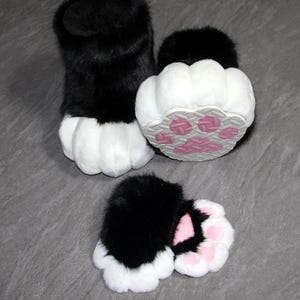 May include: Furry black and white paw-shaped costume feet. The paws have a black upper portion and white pads. The soles feature pink paw prints on a gray background. The set includes two feet and two smaller paw pads.