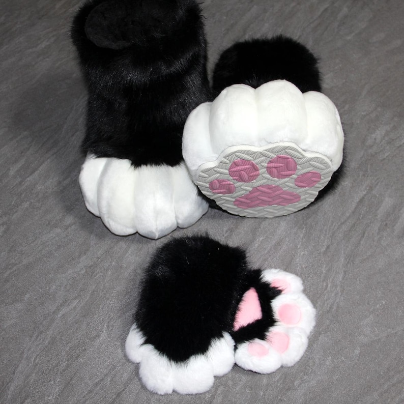 May include: Furry black and white paw-shaped costume feet. The paws have a black upper portion and white pads. The soles feature pink paw prints on a gray background. The set includes two feet and two smaller paw pads.