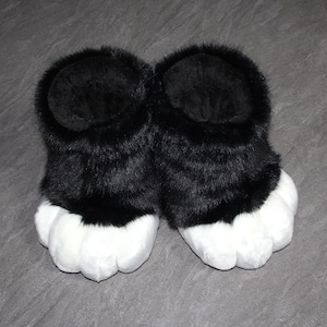 May include: A pair of black and white furry paw slippers. The slippers have a black upper portion with a fluffy texture and white, padded paw-shaped bottoms. The slippers are designed to resemble animal paws.