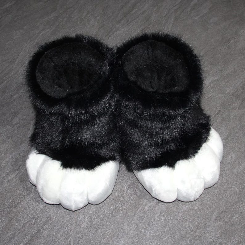 May include: A pair of black and white furry paw slippers. The slippers have a black upper portion with a fluffy texture and white, padded paw-shaped bottoms. The slippers are designed to resemble animal paws.