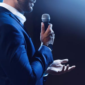 May include: A dark blue suit jacket worn over a crisp white collared shirt. A silver handheld microphone is held in one hand, while the other hand is open with palm facing upward. A brown leather watch is visible on the wrist of the hand holding the mic.