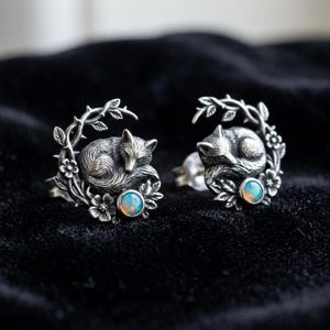 May include: A pair of silver-toned fox earrings. Each earring features a sleeping fox nestled within a floral and leaf design. A small, round, opalescent stone is set below the fox. The earrings are detailed and have a vintage aesthetic.