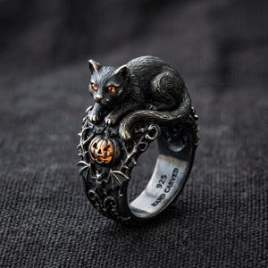 May include: A silver-toned ring featuring a black cat perched above a carved pumpkin. The ring has intricate detailing, including bats and scrollwork. The cat has orange eyes, and the ring is marked "925 HAND CARVED".