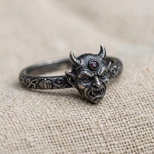 May include: A silver-toned ring featuring a detailed devil face with horns and a small, round, dark red gemstone set in the forehead. The band is decorated with an embossed pattern of swirling vines and small pumpkin shapes along the sides.