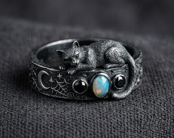 Puede incluir: Anillo de plata con una detallada figura de gato en la banda. El anillo presenta un diseño de media luna y telaraña, con tres piedras incrustadas: dos negras y una ópalo. El anillo está sobre un fondo gris oscuro.