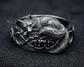 May include: A silver-toned ring featuring a detailed fox design. The fox is curled up on a decorative band with three black gemstones. The ring has an antique, gothic aesthetic.