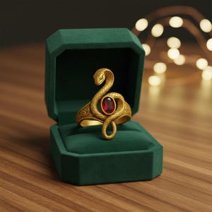Puede incluir: Anillo dorado con forma de serpiente, con una gema roja ovalada. Se muestra en un joyero de terciopelo verde oscuro abierto, sobre una superficie de madera. Joyería.
