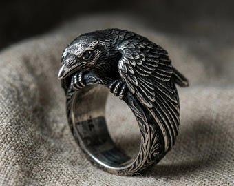 May include: A silver-toned metal ring featuring a detailed, three-dimensional raven perched on a branch. The bird has textured feathers and a dark eye, while the band of the ring is decorated with intricate, carved leaf-like patterns throughout.