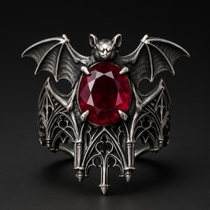 May include: A silver ring featuring a bat with outstretched wings above a large, oval, red gemstone. The ring has a gothic design with intricate detailing and a dark, antique finish. The ring is set against a black background.