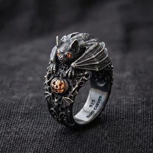 May include: A silver ring featuring a detailed bat with orange eyes perched above a small carved pumpkin. The band includes intricate filigree patterns and the text 925 HAND CARVED engraved on the inner surface of the metal ring structure.