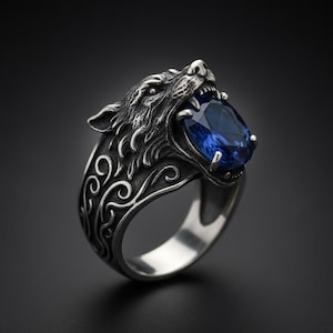 Op de afbeelding: Zilveren ring met een gedetailleerd wolfskopontwerp, met een grote, ovale, diepblauwe edelsteen. De ringband heeft een sierlijk, wervelend patroon. Een opvallend sieraad met een gedetailleerd en opvallend ontwerp.