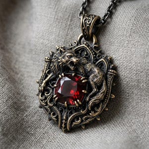 May include: A detailed pendant necklace featuring a snarling wolf design. The pendant has a dark bronze finish and a large, square, red gemstone at its center. The necklace chain is a dark, gunmetal color.