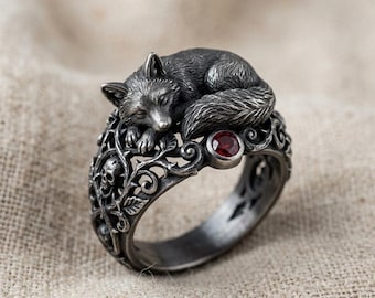 May include: A silver-toned ring featuring a sleeping fox atop an ornate band. The band has a detailed design with skulls and swirling patterns. A small, round, red gemstone is set into the ring. The fox is intricately detailed.