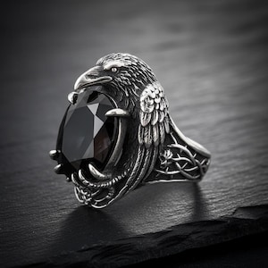 May include: A silver-colored ring featuring a detailed raven design. The raven's head and wings frame a large, black, teardrop-shaped gemstone. The ring has intricate detailing on the band, showcasing a gothic or fantasy theme.