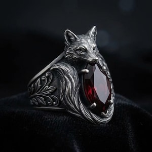 May include: A silver-toned ring featuring a fox design. The fox is sculpted with intricate detail, holding a large, teardrop-shaped, deep red gemstone. The ring has an ornate, floral-inspired band.