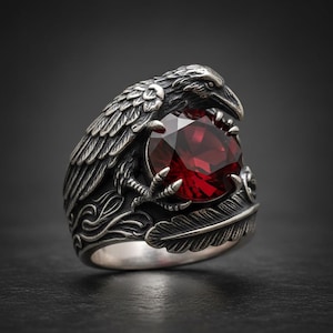 May include: A silver ring featuring a detailed raven design clutching a large, faceted, red gemstone. The ring has intricate feather and scrollwork details, creating a gothic or fantasy aesthetic. The gemstone is a deep, rich red color.