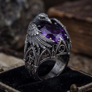 May include: A silver ring featuring a raven design with a large, oval, purple gemstone. The raven is perched atop the ring, with detailed wings and a gothic-style setting. The ring is displayed in a black velvet box.