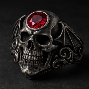 May include: A silver skull ring with a large, round, red gemstone set in the forehead. The ring features detailed carvings, including bat-like wings on the sides and ornate scrollwork. The ring is a gothic or biker style.