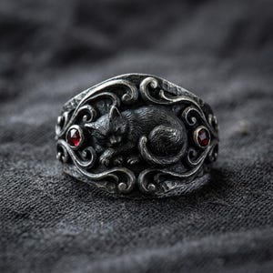 May include: A silver ring featuring a detailed, sleeping black cat design. The ring has ornate scrollwork and two small, round red gemstones. The cat is curled up in the center of the ring.