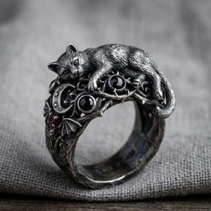 May include: A silver-toned ring featuring a detailed cat resting atop a decorative band. The band includes crescent moons, stars, and black and red gemstones. The ring has a gothic or fantasy theme.
