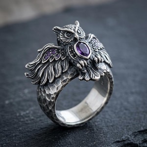 May include: A silver ring with an owl motif. The owl is adorned with a central purple gemstone and smaller purple stones on the wings. The ring has a textured band and detailed feathering.