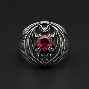 May include: A silver ring featuring a bat design with outstretched wings, centered around a large, oval-cut, red gemstone. The ring has a gothic style with intricate detailing and is set against a dark background.