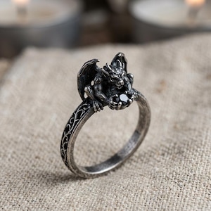 May include: A silver-toned ring featuring a detailed gargoyle figure perched on the band. The gargoyle holds a small, round, faceted black stone in its hands. The band of the ring is decorated with an intricate, embossed vine-like pattern.