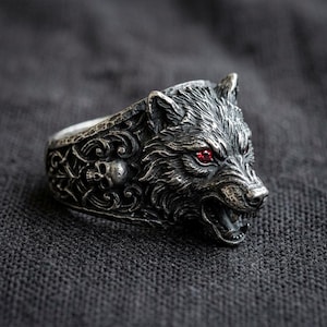 Puede incluir: Un anillo de metal plateado que presenta una cabeza de lobo detallada con dos pequeñas piedras rojas incrustadas como ojos. La banda está decorada con intrincados patrones de volutas y una pequeña calavera humana esculpida en el lateral.