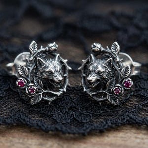 Könnte beinhalten: Ein Paar silberfarbene Ohrstecker. Jeder Ohrring zeigt einen detaillierten Wolfskopf, umgeben von einem Kranz aus Blättern, Rosen und Dornen. Kleine, runde, rote Edelsteine akzentuieren das Design.