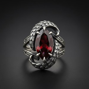 Könnte beinhalten: Ein silberner Ring mit einem großen, tropfenförmigen, tiefroten Edelstein. Der Ring hat aufwendige flügelartige Designs auf beiden Seiten des Edelsteins mit detaillierten Federtexturen. Der Ring ist vor einem dunklen Hintergrund platziert.