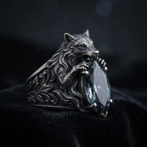 May include: A silver-toned ring featuring a detailed wolf design biting a large, light blue gemstone. The ring has intricate floral patterns on the band, creating a gothic or fantasy aesthetic. The gemstone is a marquise cut.