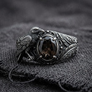 May include: An oxidized silver ring featuring a sculpted raven perched beside an oval-cut dark brown gemstone. The band is detailed with intricate feather engravings and ornate scrollwork surrounding the central stone setting.
