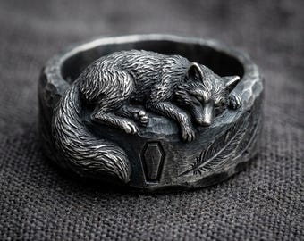 May include: A silver ring featuring a detailed sleeping fox design. The ring has a textured band with carved details including a coffin and feather. The fox is lying down, with its eyes closed, and its tail curled around the band.