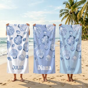 May include: Three rectangular beach towels featuring a repeating pattern of blue and white seashells. The left towel is white with the name Julia, the centre is light blue with Diana, and the right is pale blue with the name Alina printed at the bottom.