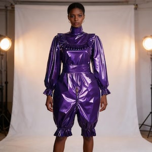 May include: A shiny, purple jumpsuit with a high neckline, long sleeves, and ruffled trim. The jumpsuit features a cinched waist and a zipper down the front. The bottom portion is a short, puffy style with ruffled cuffs. The outfit is made of a glossy material.